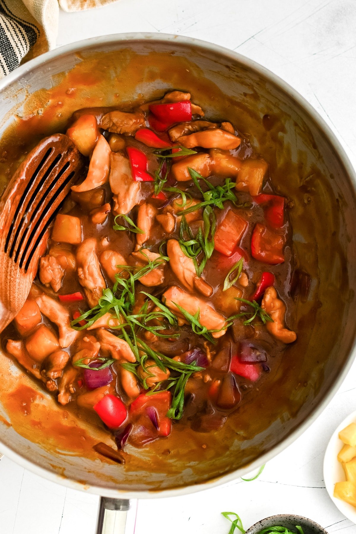 Pineapple chicken simmering in a skillet with chicken, red bell peppers, onions, pineapple chunks, and green onions on top.