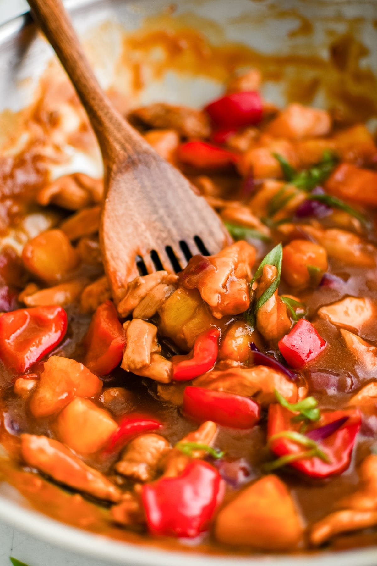 Close-up of pineapple chicken being stirred with a wooden spoon, showing chunks of chicken, pineapple, and bell pepper coated in sauce.