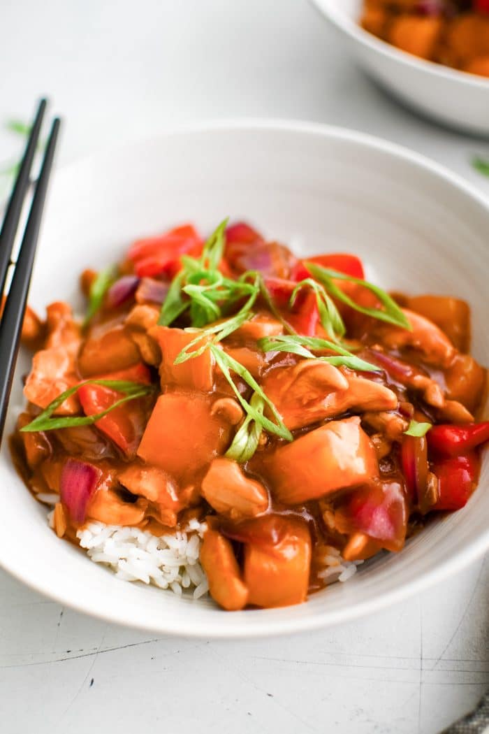 Bowl of pineapple chicken served over white rice, garnished with green onions and filled with chicken, pineapple, red peppers, and onions.