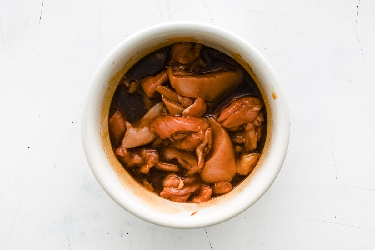 Raw chicken pieces marinating in a dark pineapple-hoisin mixture inside a white bowl.