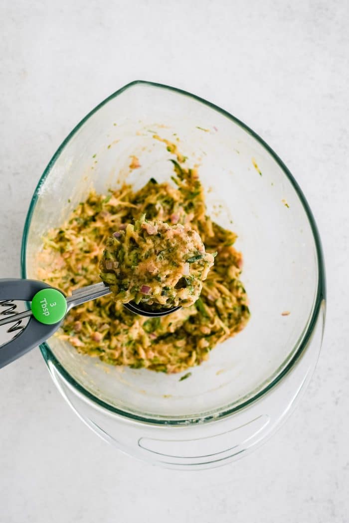 Cookie scoop removing a scoopful of zucchini fritter mixture from a large glass mixing bowl.