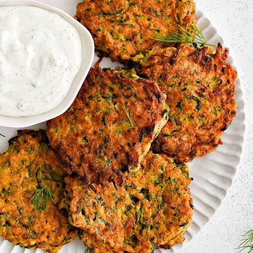 Large white plate filled with six crispy pan-fried zucchini fritters with a side of herbed yogurt dip in a small white serving bowl.