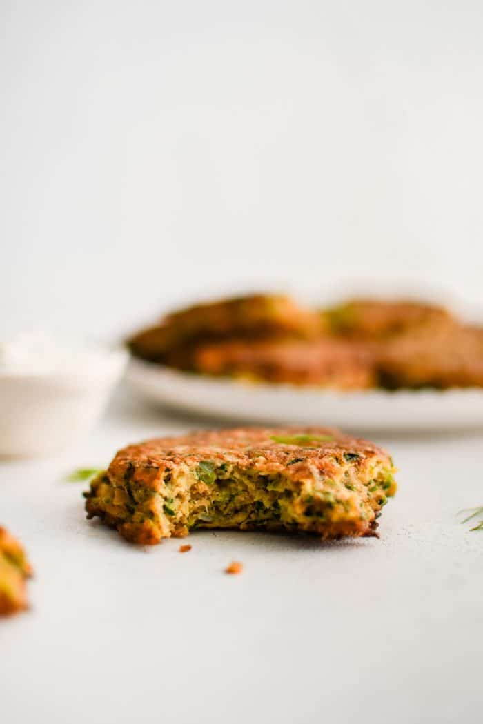 A single zucchini fritter with a bite removed set in front of a large plate filled with the remaining golden, crispy fritters.