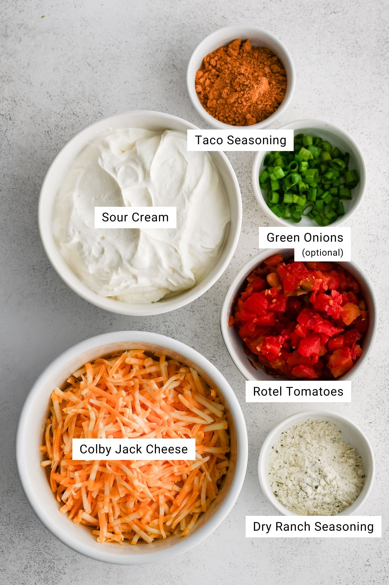 Overhead photo of the six ingredients needed to make TikTok-famous boat dip, including bowls of sour cream, shredded Colby Jack cheese, Rotel tomatoes, taco seasoning, dry ranch seasoning, and optional sliced green onions. 