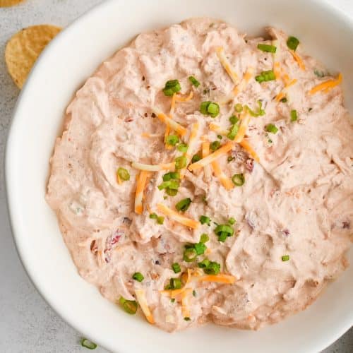 Boat dip made with shredded cheddar cheese, green onions, sour cream, drained rotel tomatoes, taco seasoning, and ranch seasoning in a large white serving bowl and garnished with additional shredded cheese and green onions.