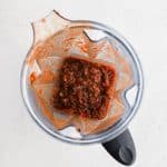 The bowl of a high speed blender filled with the leftover solids from strained chile colorado sauce with half a cup of chicken broth.