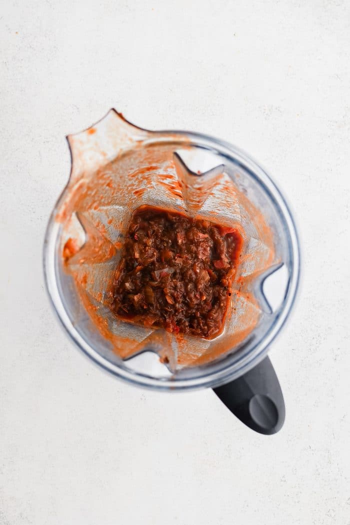 The bowl of a high speed blender filled with the leftover solids from strained chile colorado sauce with half a cup of chicken broth.