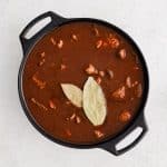 Large cast iron pot filled with seared beef chunks covered in homemade chile colorado sauce and topped with two bay leaves.