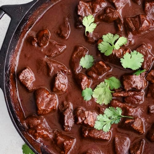 Homemade chile colorado with chunks of beef in a large cast iron pot and garnished with fresh cilantro.