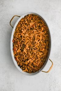 Large oval-shaped casserole dish filled with the cheesy, meat, bean, corn, and veggie filling for cowboy casserole in an even layer and topped with shredded cheddar cheese.