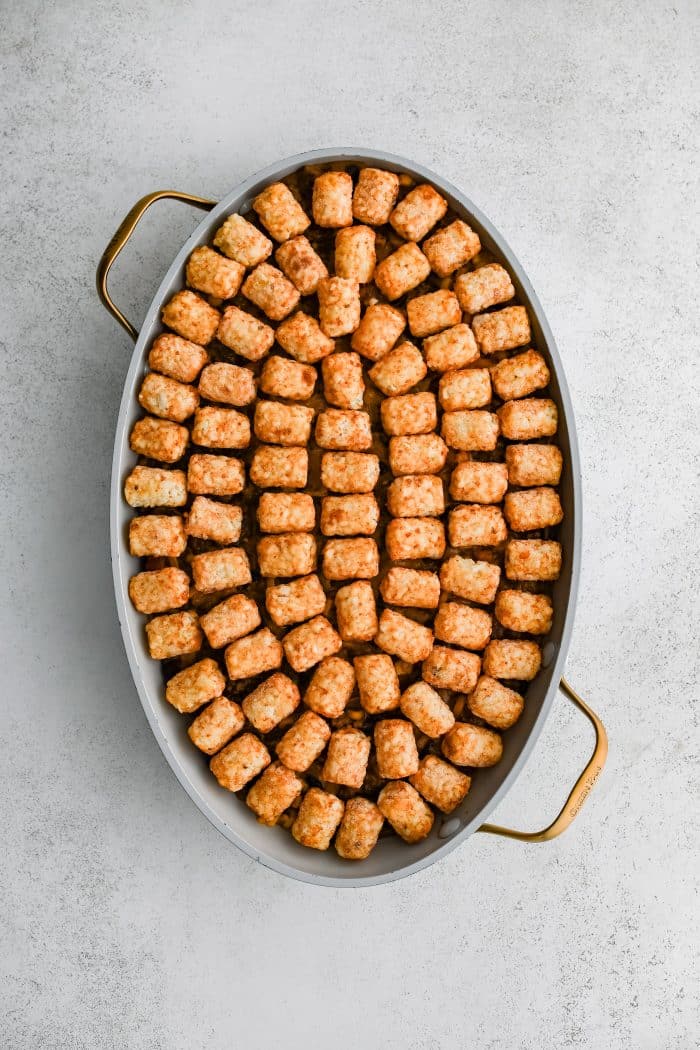 A single layer of arranged frozen tater tots placed on top of a large oval-shaped casserole dish filled with the cheesy, meat, bean, corn, and veggie filling for cowboy casserole in an even layer and topped with shredded cheddar cheese.