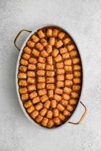 A single layer of arranged crispy baked tater tots placed in a large oval-shaped casserole dish filled with the cheesy, meat, bean, corn, and veggie filling for cowboy casserole in an even layer and topped with shredded cheddar cheese.