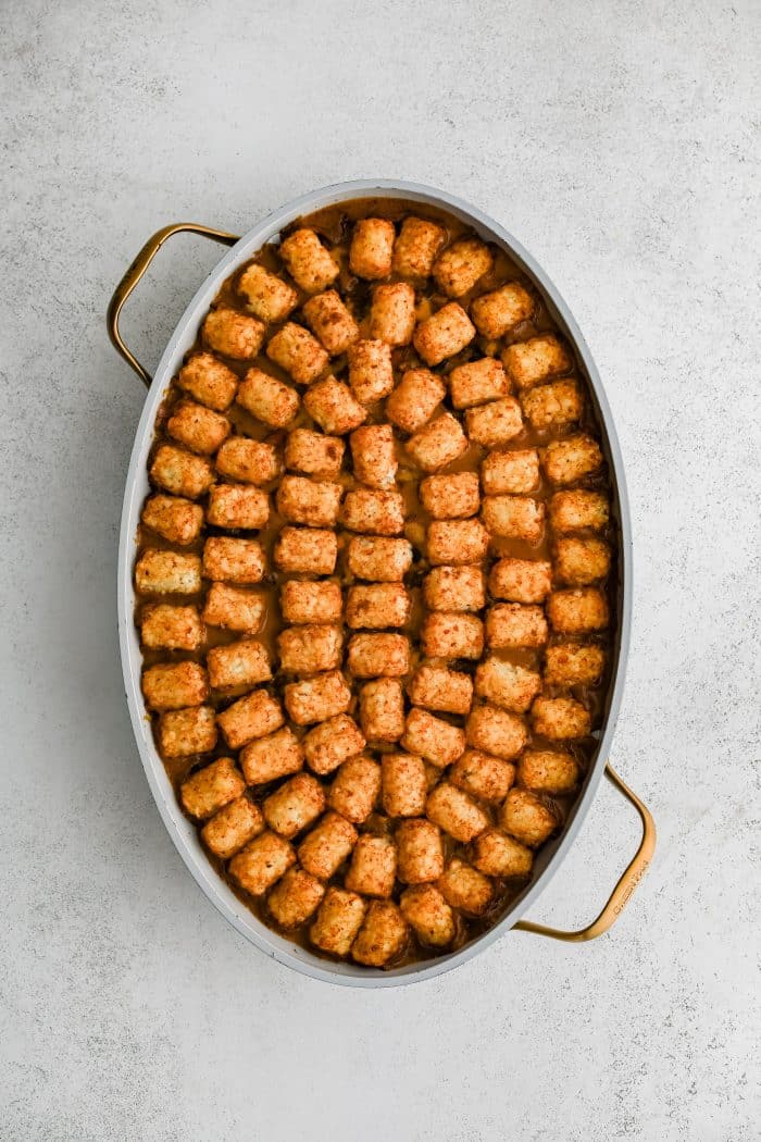 A single layer of arranged crispy baked tater tots placed in a large oval-shaped casserole dish filled with the cheesy, meat, bean, corn, and veggie filling for cowboy casserole in an even layer and topped with shredded cheddar cheese.