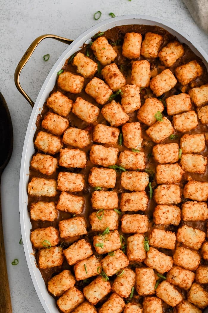 A single layer of arranged crispy baked tater tots placed in a large oval-shaped casserole dish filled with the cheesy, meat, bean, corn, and veggie filling for cowboy casserole in an even layer and topped with shredded cheddar cheese.