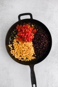 Canned tomatoes, canned corn, and canned black beans added to a large cast iron skillet filled with softened green onions and green bell pepper.