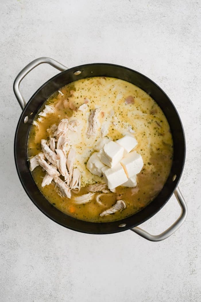 Shredded chicken, cubed cream cheese, and heavy cream added to a large soup pot filled with simmering pasta, bacon. and veggies in broth.