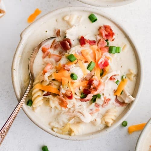 Three white soup bowls filled with super creamy crack chicken soup filled with rotini pasta, shredded chicken, bacon, and topped with shredded cheddar cheese, chopped green onions, and even more crispy cooked bacon bits.