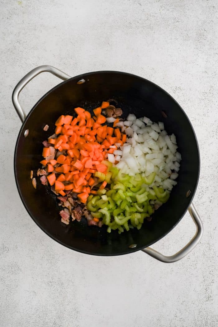 Large soup pot filled with crispy cooked bacon bits covered by diced carrots, celery, and onions.