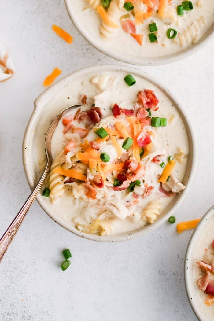 Three white soup bowls filled with super creamy crack chicken soup filled with rotini pasta, shredded chicken, bacon, and topped with shredded cheddar cheese, chopped green onions, and even more crispy cooked bacon bits.