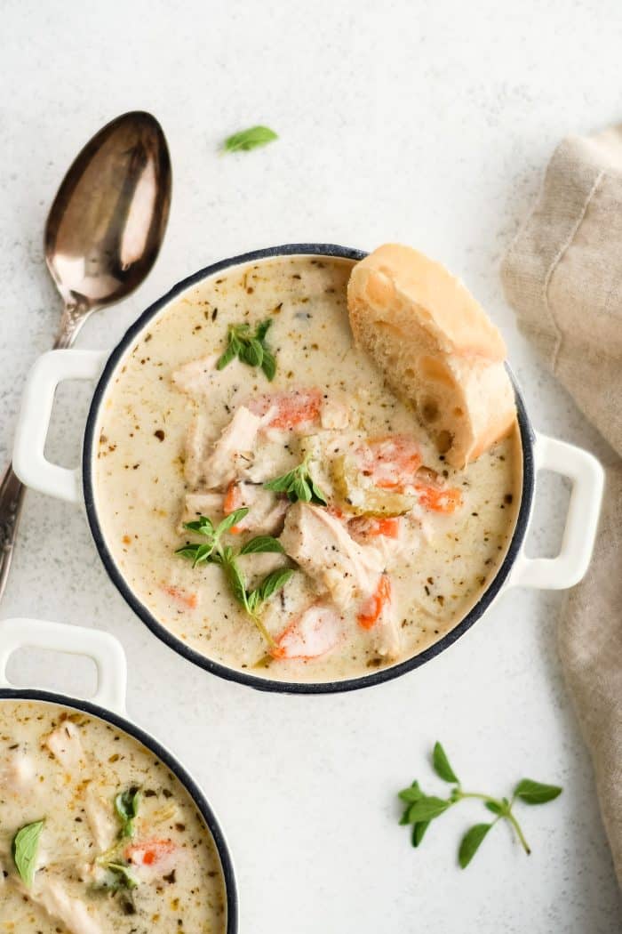 Two small white soup bowl filled with creamy chicken soup garnished with fresh parsley and oregano and topped with a single slice of fresh baked bread.