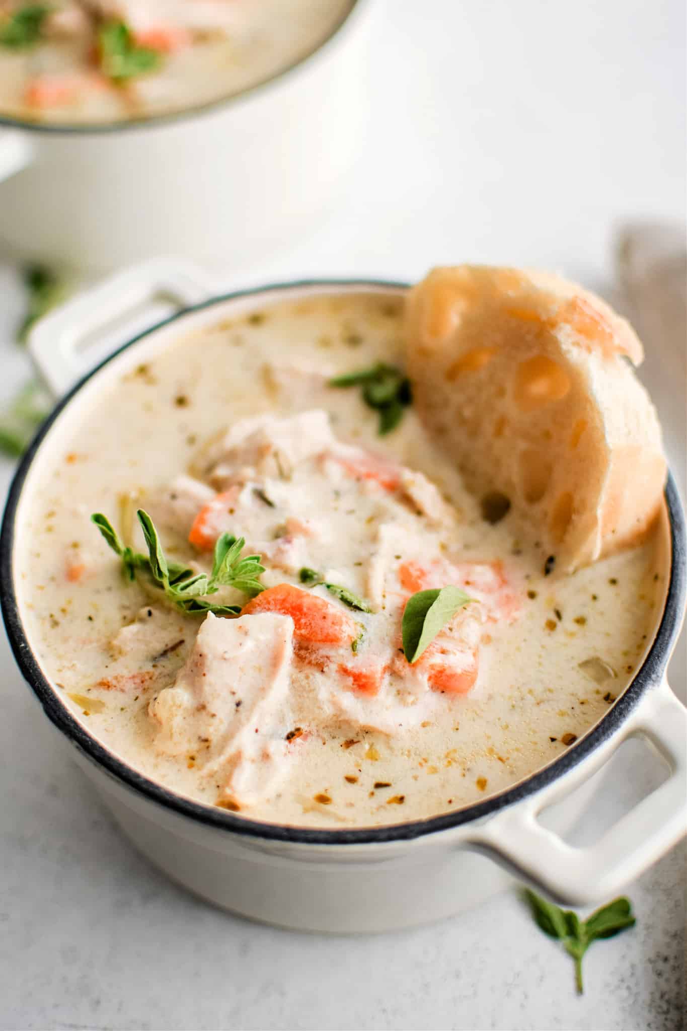 Small white soup bowl filled with creamy chicken soup garnished with fresh parsley and oregano and topped with a single slice of fresh baked bread.