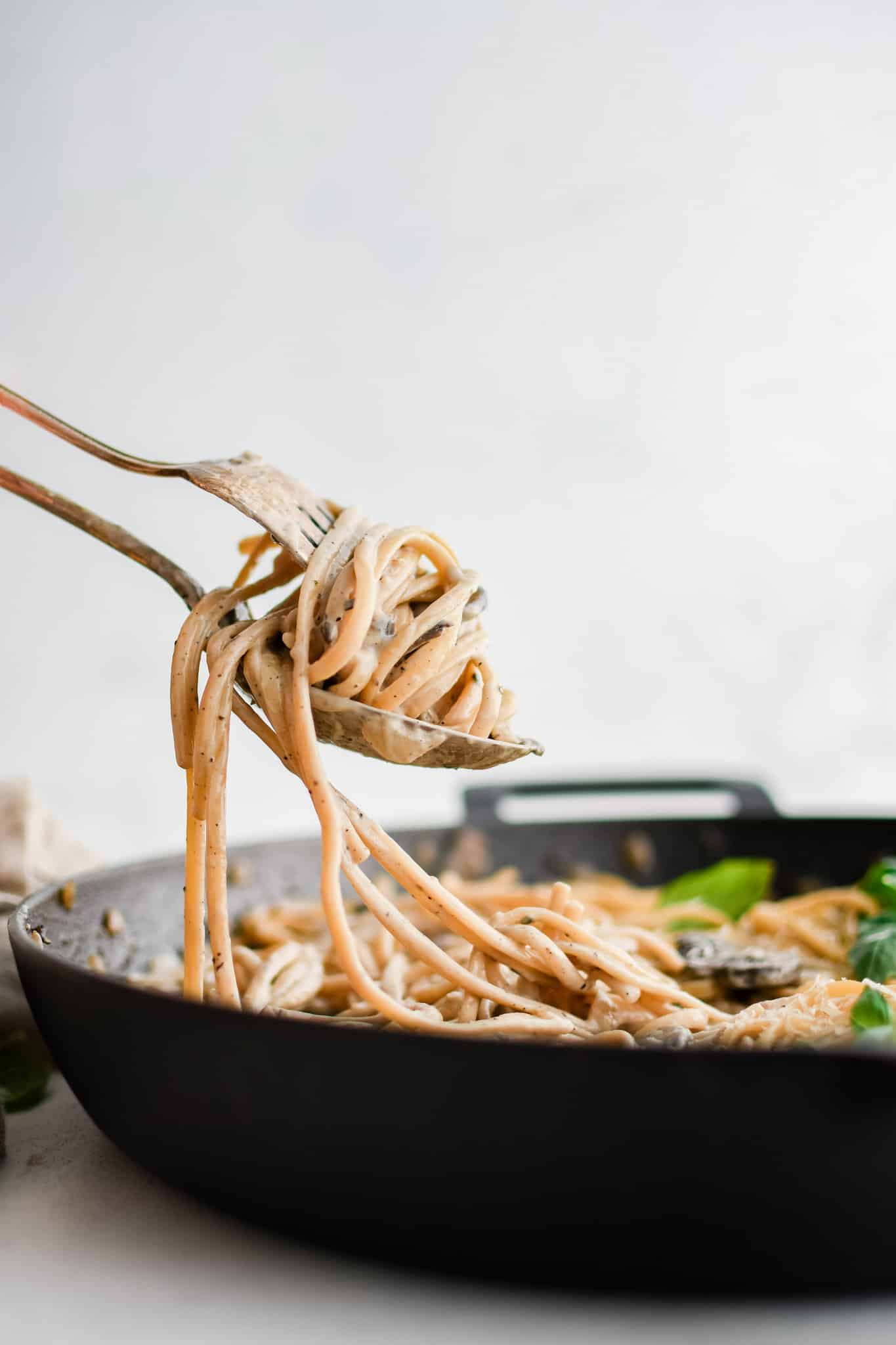 Twisted creamy linguine pasta around a fork and spoon hovering over a large cast iron skillet filled with creamy mushroom pasta.