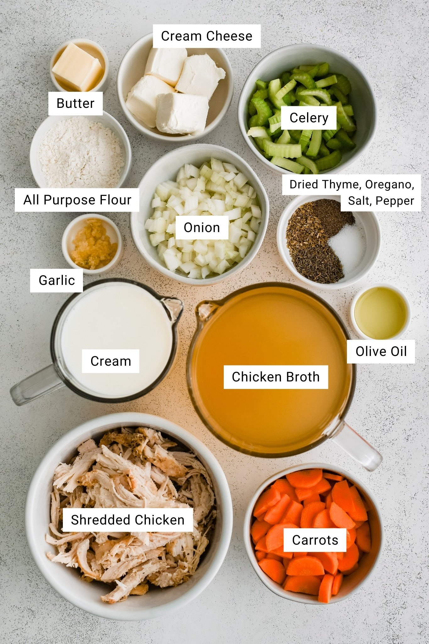 Overhead image showing labeled ingredients for creamy chicken soup, including cream cheese, celery, onion, carrots, shredded chicken, garlic, all-purpose flour, butter, olive oil, cream, chicken broth, and dried seasonings.