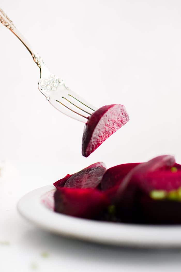 Single sliced beet one a fork hovering above a white plate filled with cooked, peeled, and sliced red beets garnished with chopped parsley.