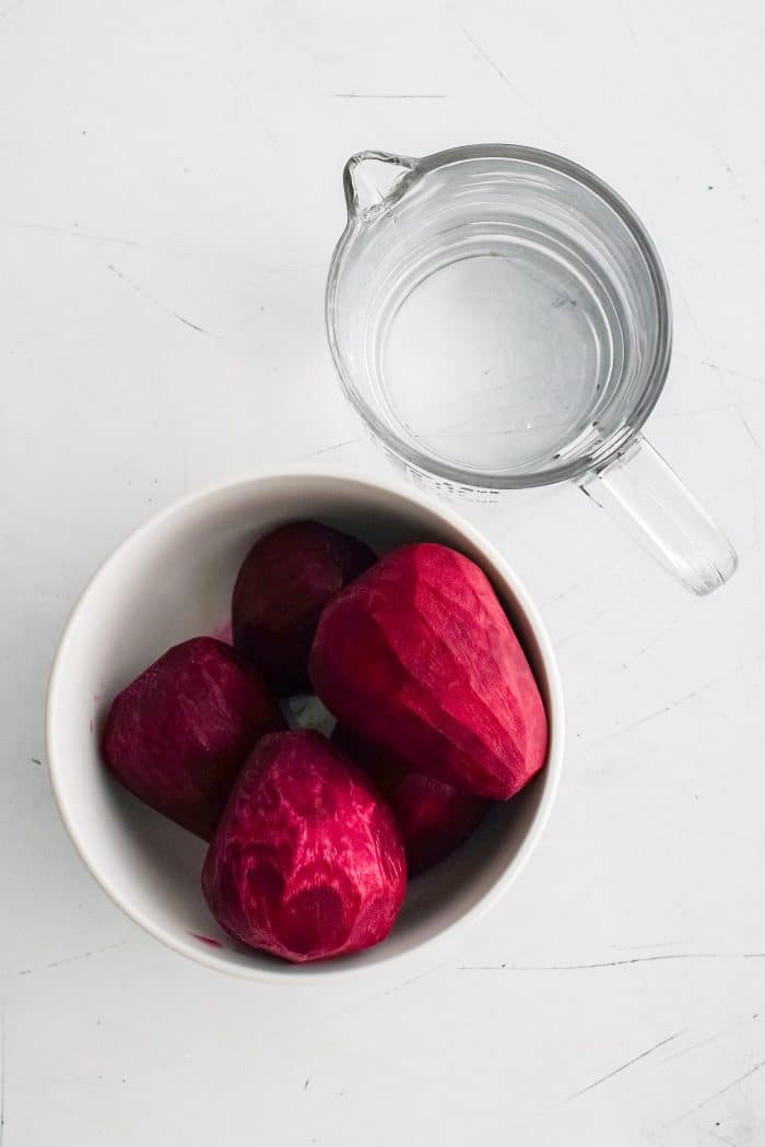 Peeled raw red beets in a white bowl next to a glass measuring cup filled with one cup of water.