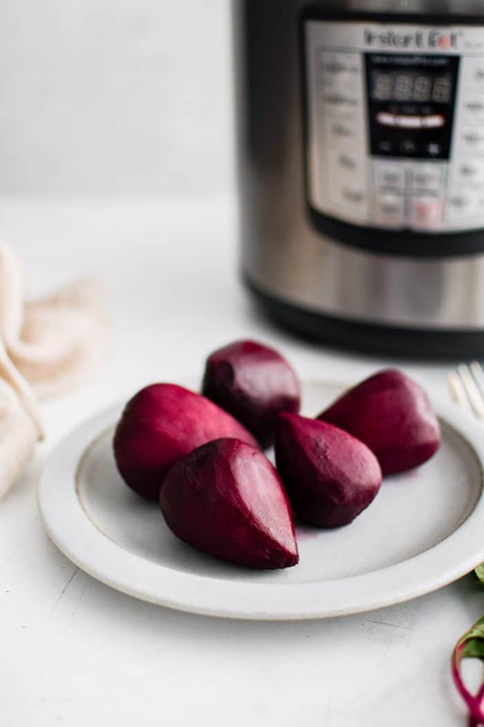 Five medium-sized cooked and peeled red beets on a white plate with the Instant Pot machine behind it.