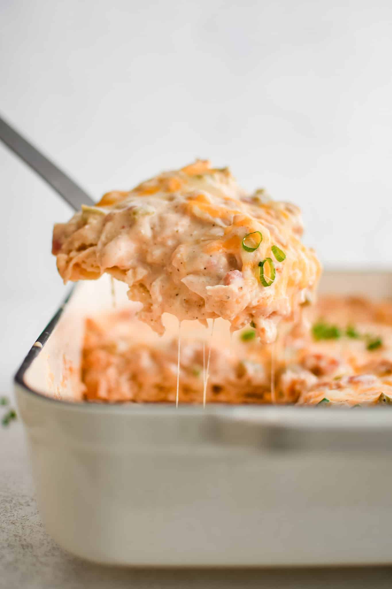 Large metal spatula scooping a large serving of hot and cheese King Ranch chicken casserole from a large white baking dish.