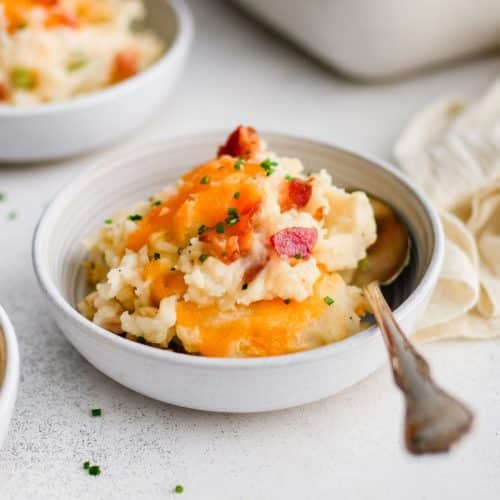 Small white serving bowl filled with loaded mashed potato casserole with melted cheddar cheese, chopped bacon bits, and garnished with minced parsley.