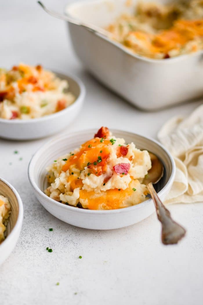 Small white serving bowl filled with loaded mashed potato casserole with melted cheddar cheese, chopped bacon bits, and garnished with minced parsley.