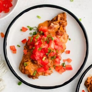 Cooked chicken breast smothered in bbq sauce, bacon bits, and melted cheddar cheese and topped with fresh chopped tomatoes and green onion on a white dinner plate.