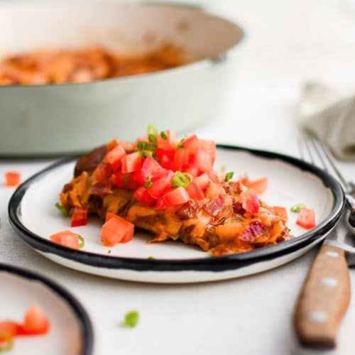 Cooked chicken breast smothered in bbq sauce, bacon bits, and melted cheddar cheese and topped with fresh chopped tomatoes and green onion on a white dinner plate.