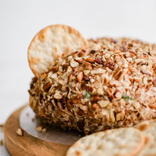 Pineapple cheese ball in the shape of a disc and coated in pecans on a small wood cutting board with a single cracker stuck in the center.