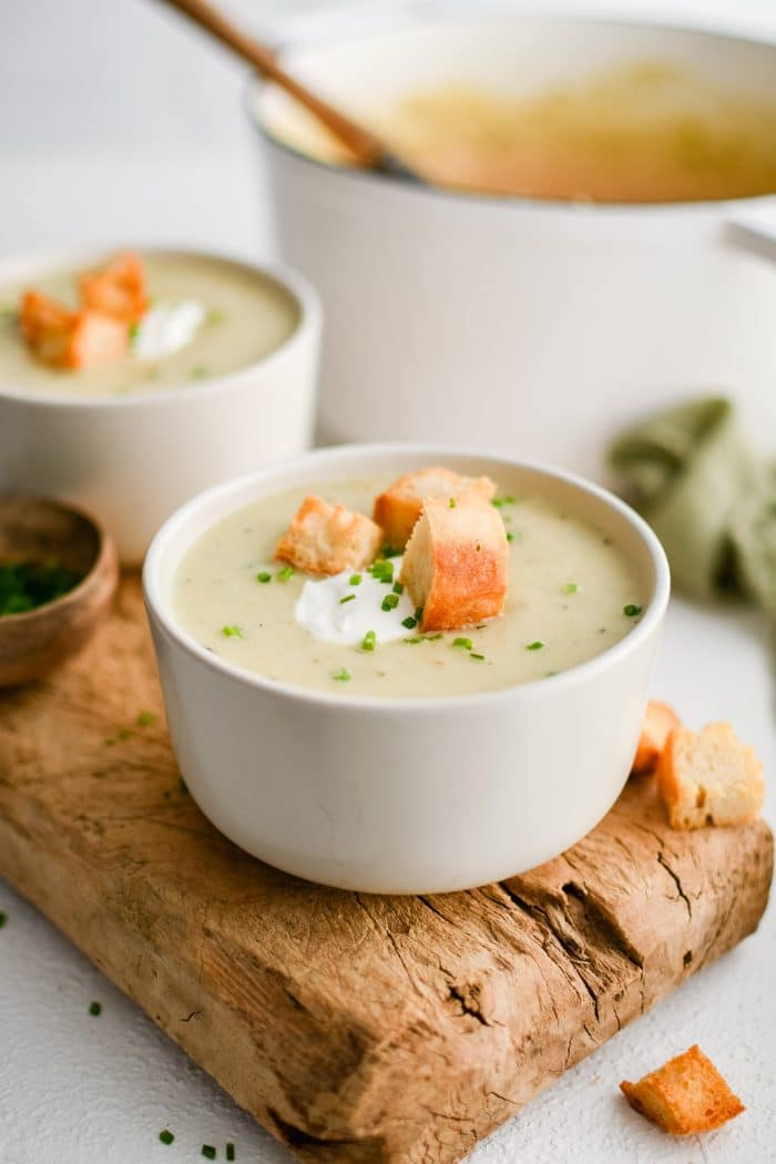 White soup bowl on a rustic wood board filled with homemade creamy potato leek soup and garnished with sour cream, homemade croutons, and freshly chopped chives.