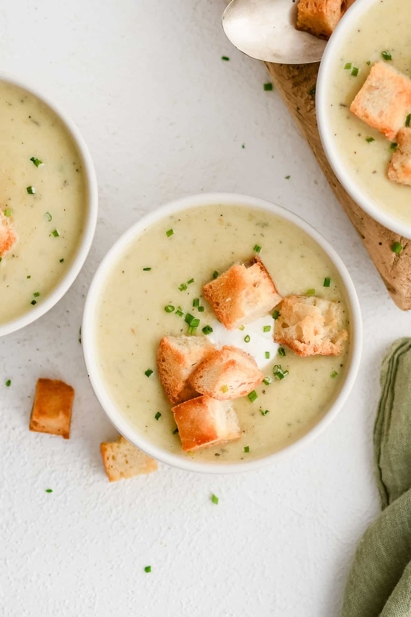 Three white bowls filled with homemade creamy potato leek soup and garnished with sour cream, homemade croutons, and freshly chopped chives.