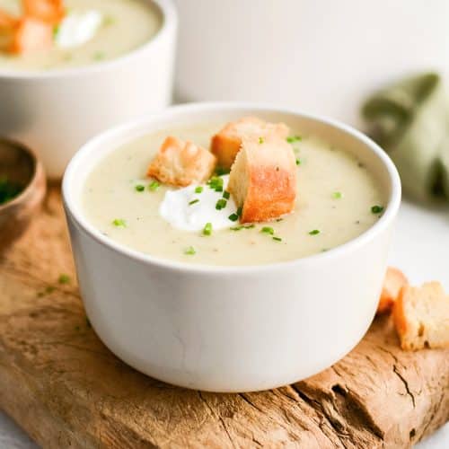 White soup bowl on a rustic wood board filled with homemade creamy potato leek soup and garnished with sour cream, homemade croutons, and freshly chopped chives.