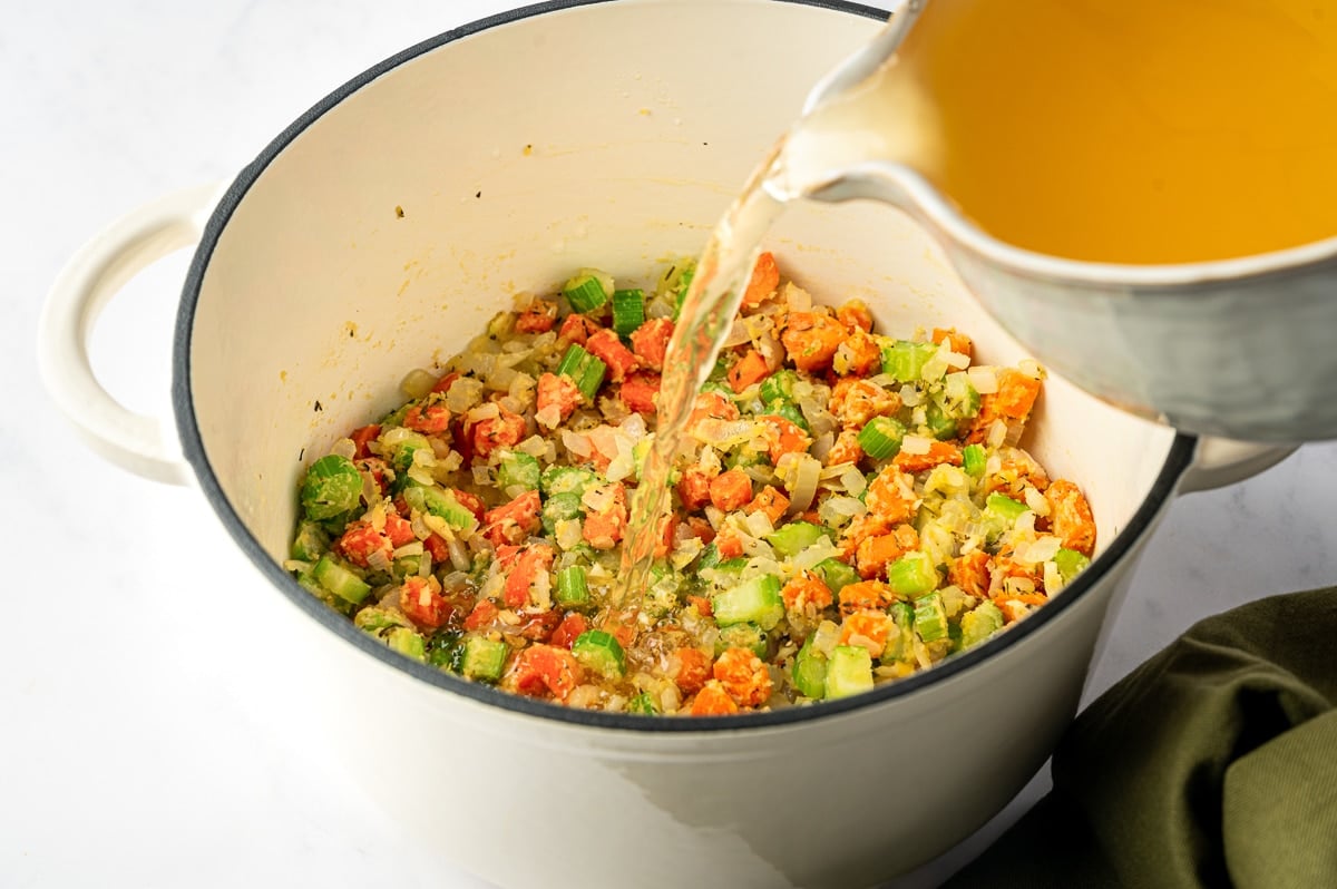 Golden chicken stock being poured into a Dutch oven filled with flour-coated carrots, celery, and onions.