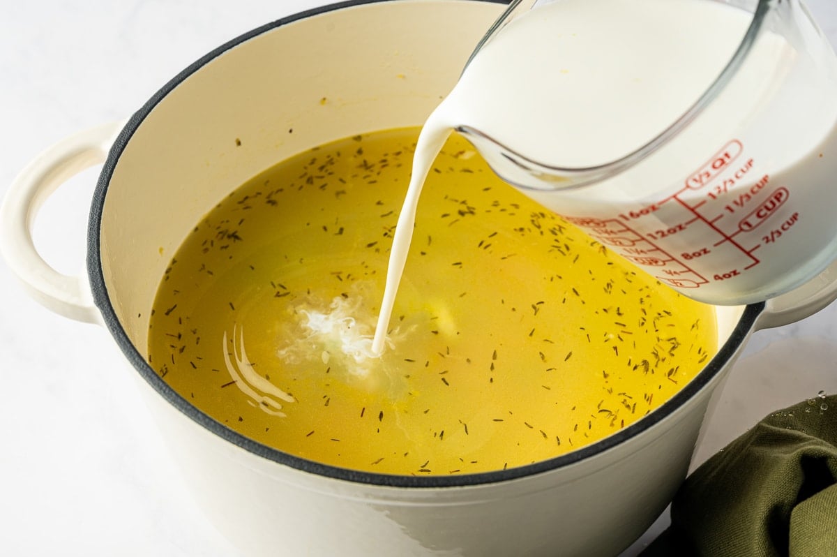 Half-and-half being poured from a glass measuring cup into a Dutch oven of golden chicken broth with thyme.