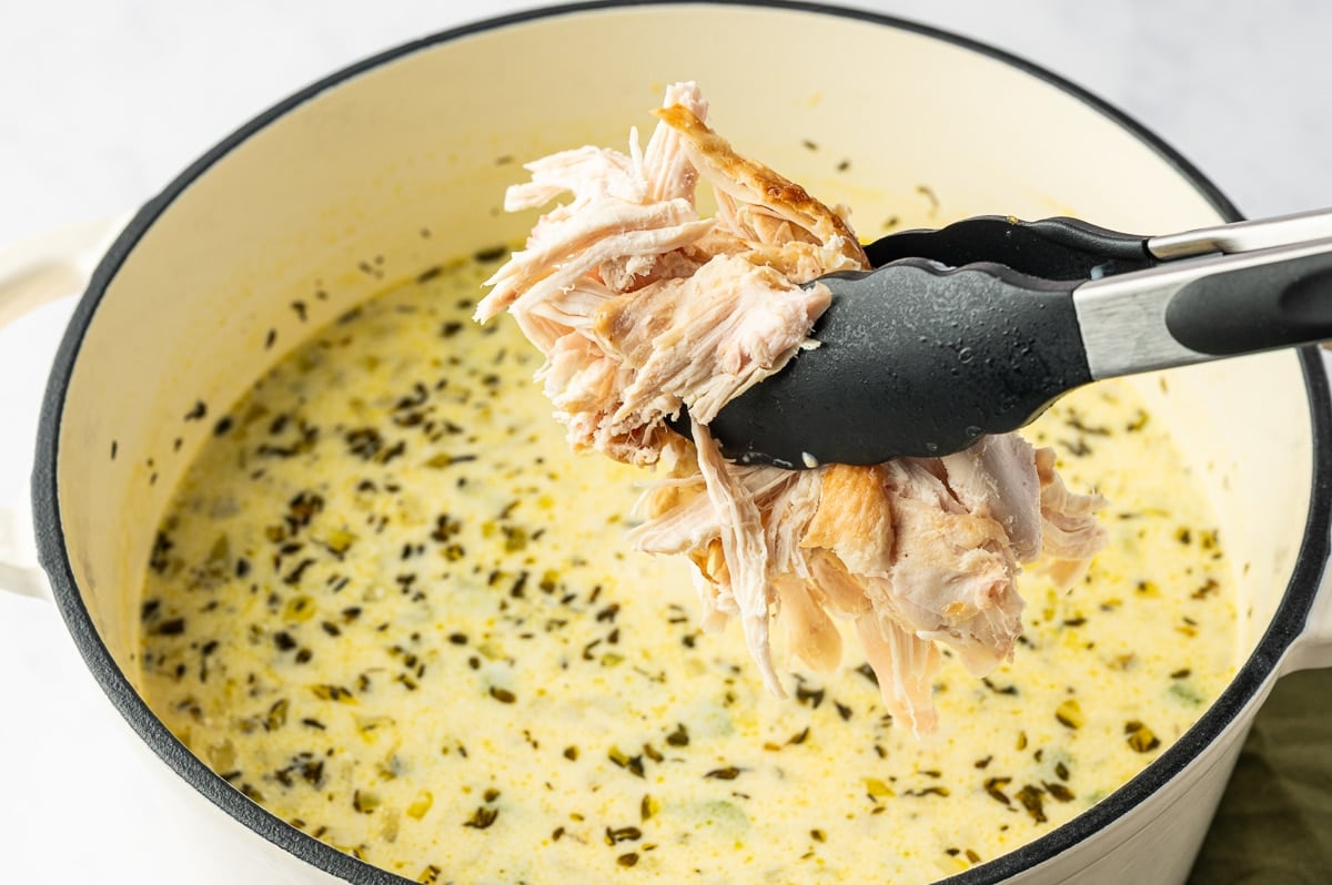 Shredded rotisserie chicken held in black kitchen tongs above a Dutch oven filled with creamy chicken soup.