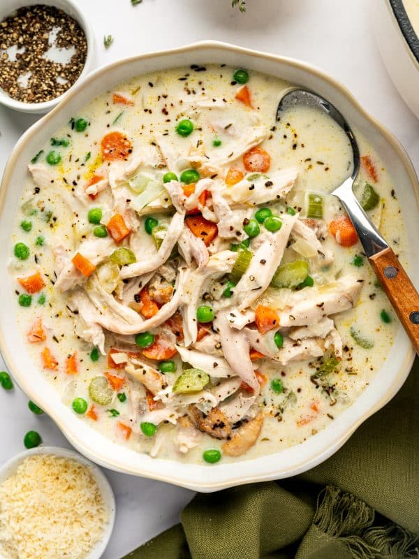 Homemade creamy chicken soup with shredded chicken, green peas, diced carrots, celery, and herbs in a white bowl with a serving spoon.