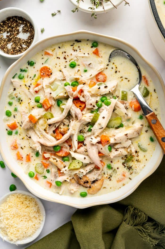 Homemade creamy chicken soup with shredded chicken, green peas, diced carrots, celery, and herbs in a white bowl with a serving spoon.