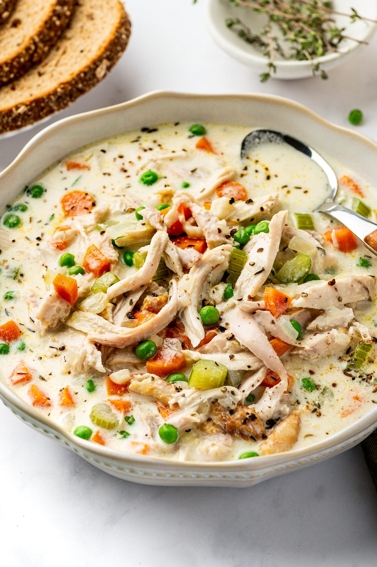 A bowl of creamy chicken soup filled with shredded rotisserie chicken, carrots, celery, and green peas, with a spoon and slices of bread nearby.