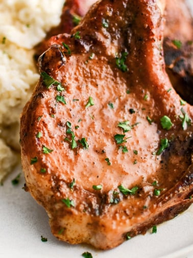 Two beautifully seasoned bone-in pork chops tilted against a pile of mashed potatoes on a white serving plate garnished with chopped parsley.