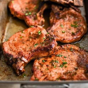 Large stainless steel baking dish filled with five oven-baked pork chops golden and crisp around the edges and garnished with freshly minced parsley.