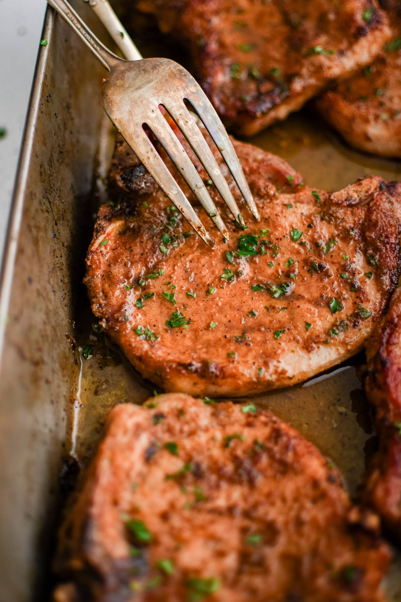 Cooked bone-in pork chop stabbed with a dinner fork to be removed from a large stainless steel baking dish.