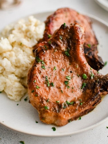 Two beautifully seasoned bone-in pork chops tilted against a pile of mashed potatoes on a white serving plate garnished with chopped parsley.