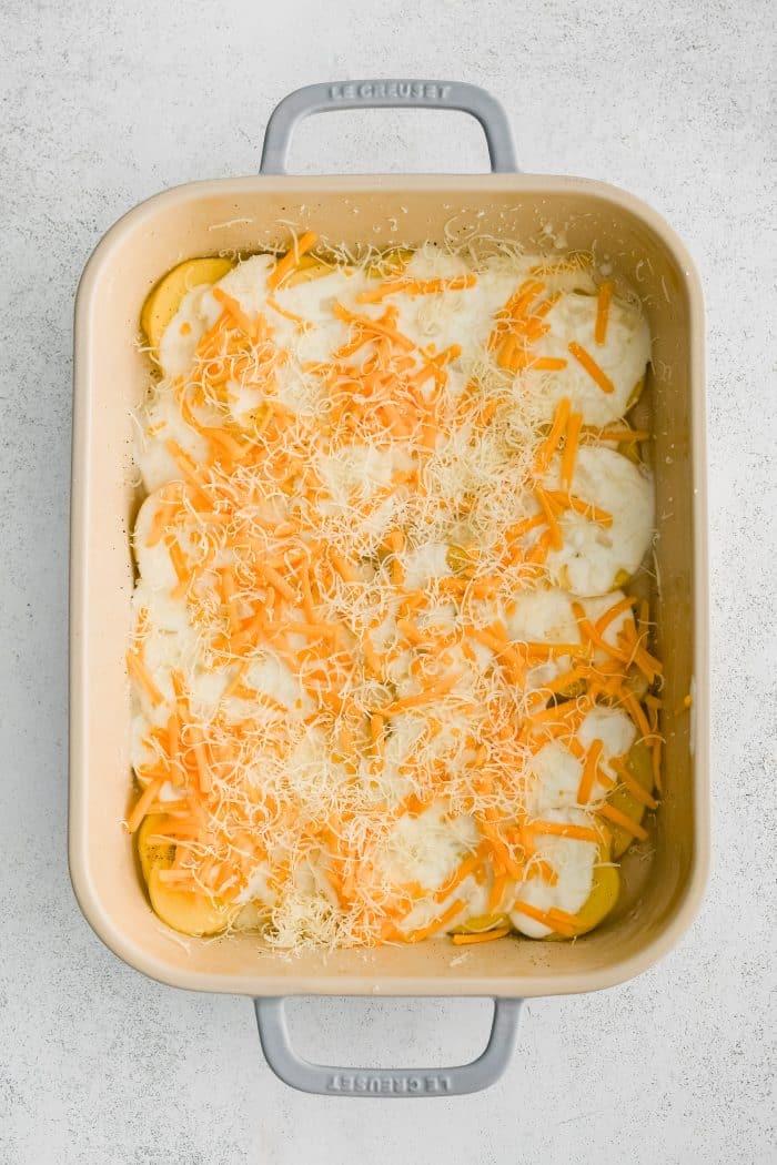 Large 9x13-inch baking dish with a bottom layer of thinly sliced potatoes topped with a middle layer of homemade cream sauce, and a top layer of shredded cheddar and parmesan cheese.